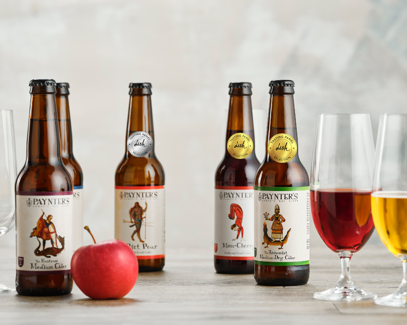 Five bottles of Paynter's cider with apples and glasses on a wooden surface.