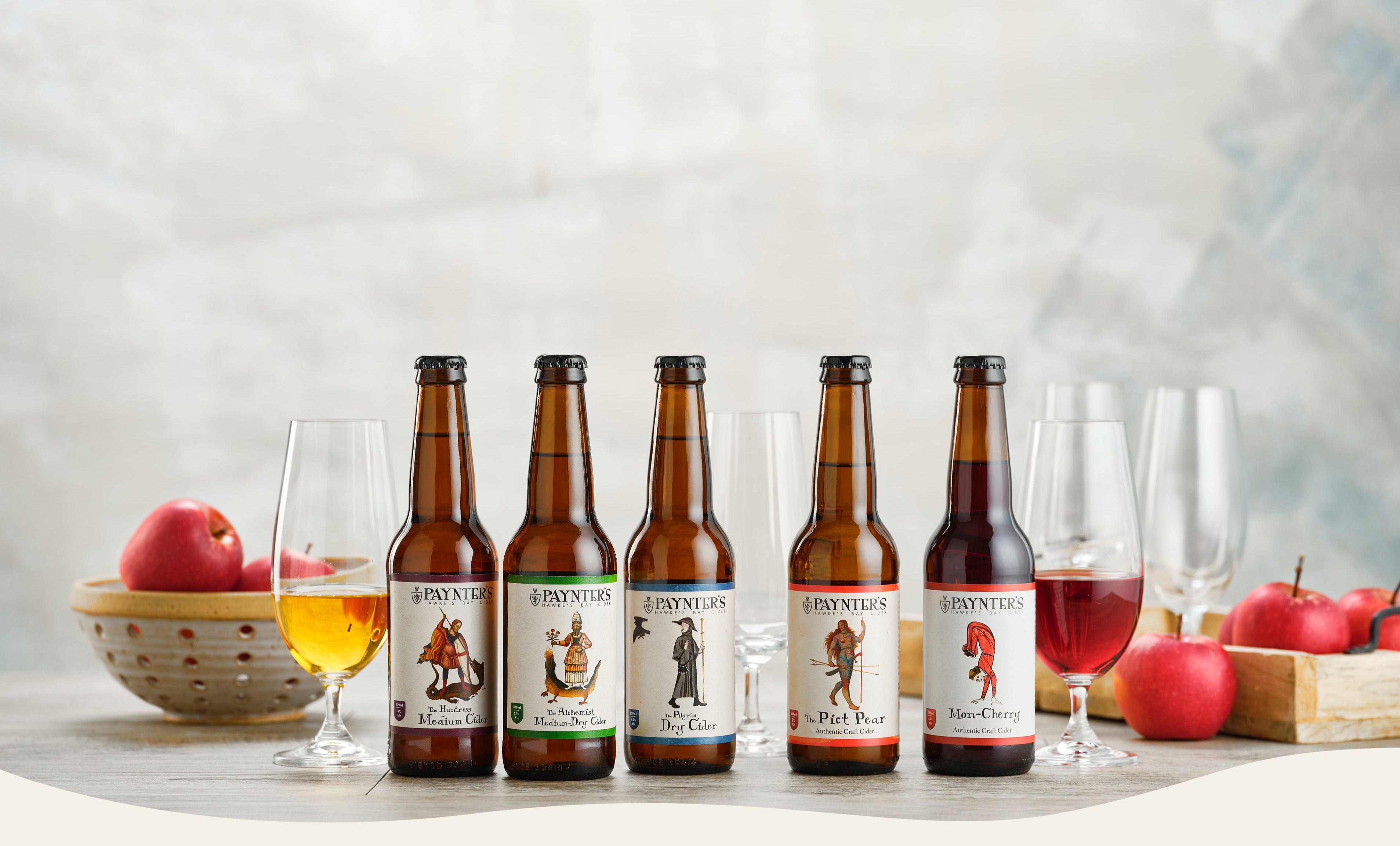 Row of Paynter's Cider brown glass bottles with labels on a table with apples and glasses.