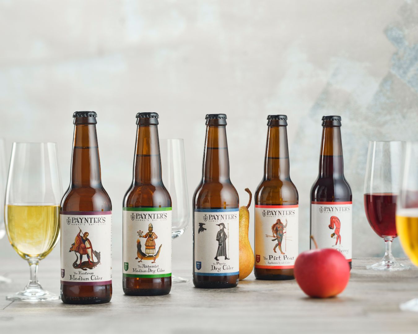 Five bottles of Paynter's beer with their labels displayed on a table with glasses and an apple.