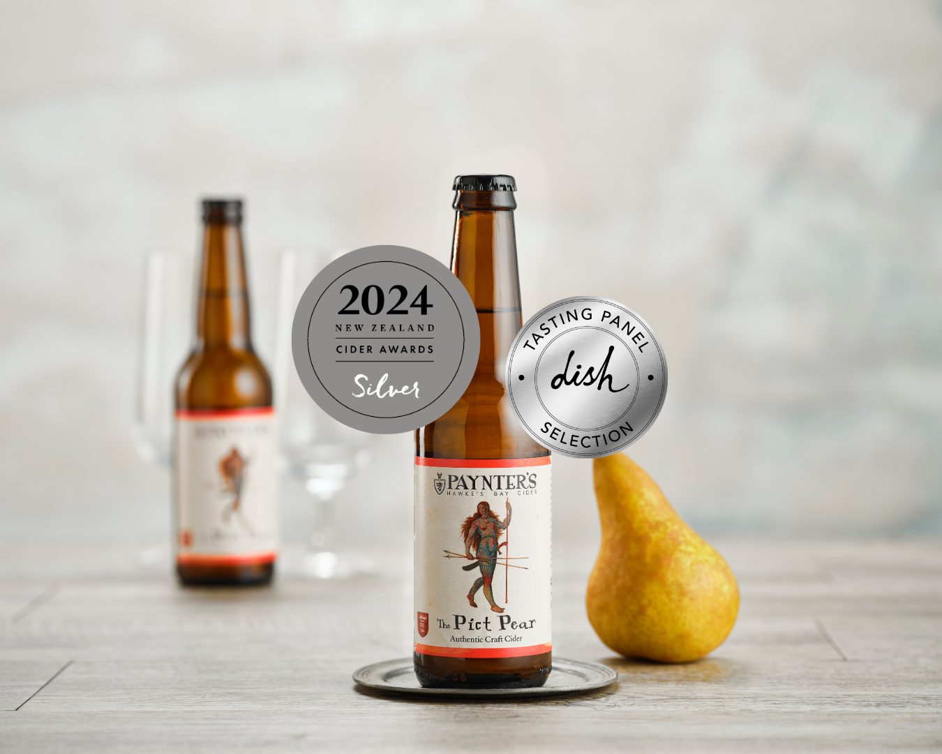 Bottle of Paynter's The Pick Pear cider with a pear on a wooden surface, featuring awards on the label.
