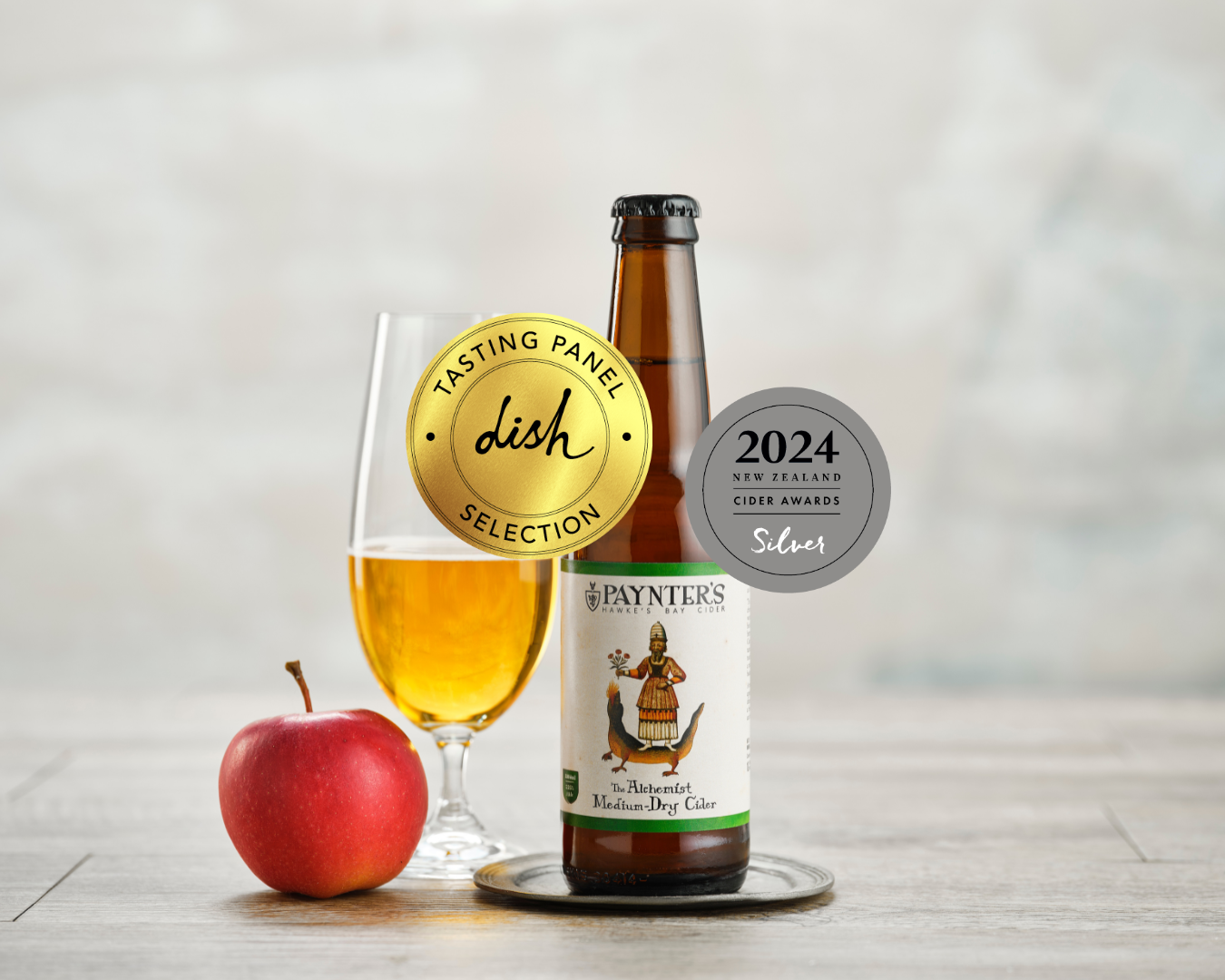 Paynter's cider bottle with a glass of cider, award stickers (gold and silver) and an apple on a wooden surface.