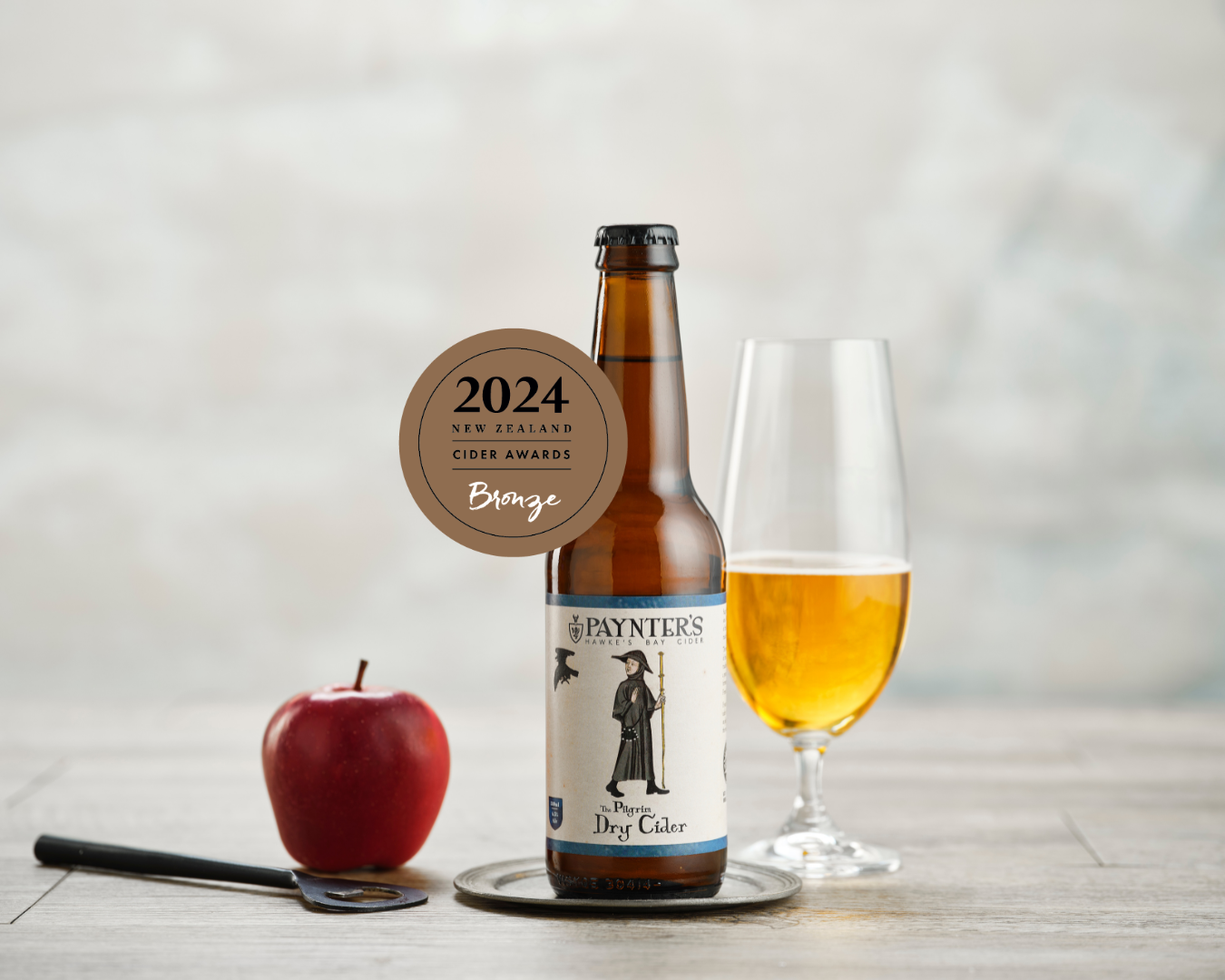 Paynter's cider bottle with a bronze 2024 Cider Awards medal sticker, a glass of cider and an apple on a wooden surface.